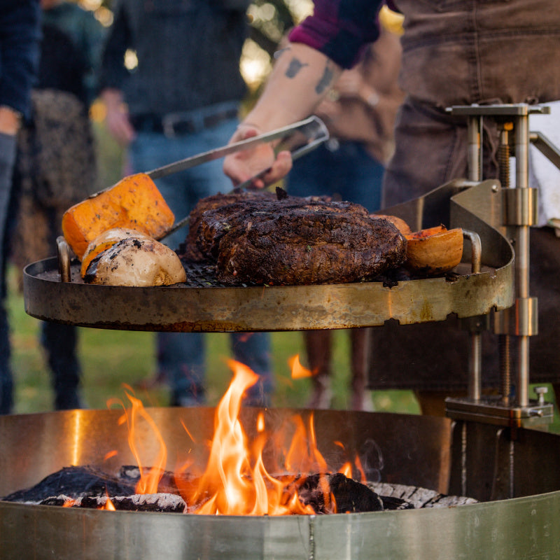 Close-up of live cook over open flame with LUX Pro Precision grill. Steaks and potatoes grilling on the Versa Grate while chef flips potatoes with LUX tongs.