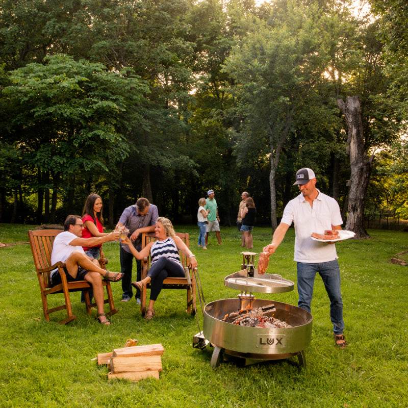 Smiling friends gathered around Lux Precision grill on sunny day in backyard. Chef grilling steaks and friends clinking glasses of wine.