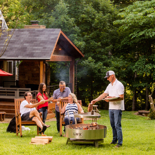 Smiling friends gathered around Lux Precision grill on sunny day in backyard. Chef grilling steaks and friends clinking glasses of wine.