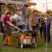 Friends gathering in backyard around LUX Pro Precision grill under strings of lights. Chef cooking meats and vegetables over open fire while guests enjoy conversation.